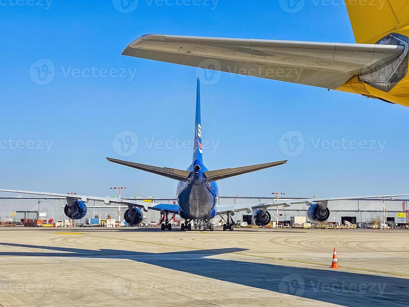Blue passenger jet tail, seen from behind, parked on tarmac with yellow wing above. High quality photo