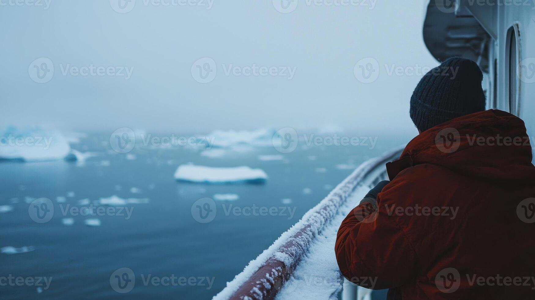 A person wearing a warm jacket stands on a ship deck, gazing at a serene icy ocean filled with floating icebergs. The cold, foggy atmosphere envelops the scene photo