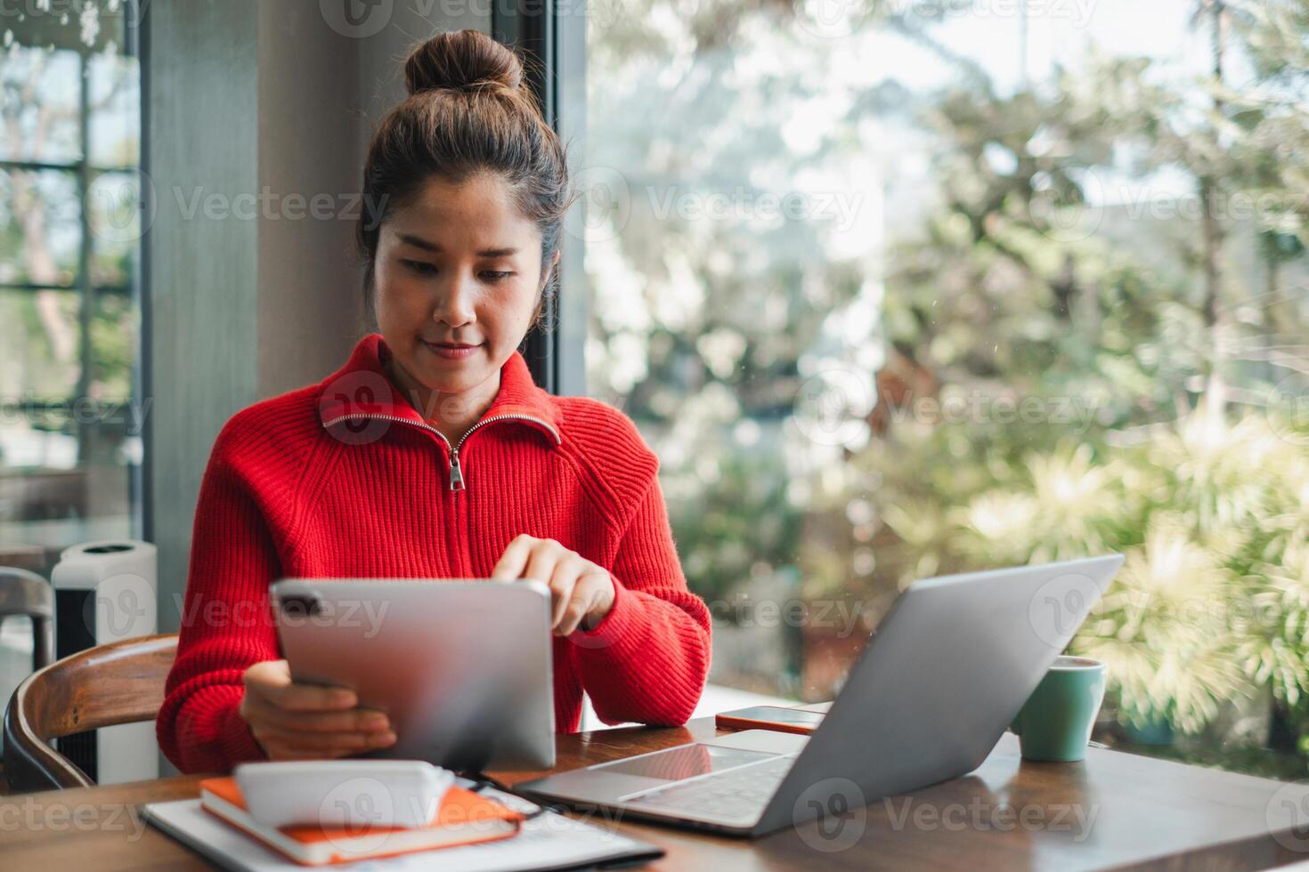 A young woman in a red sweater uses a tablet and laptop at a wooden desk in a bright, modern office with large windows. photo