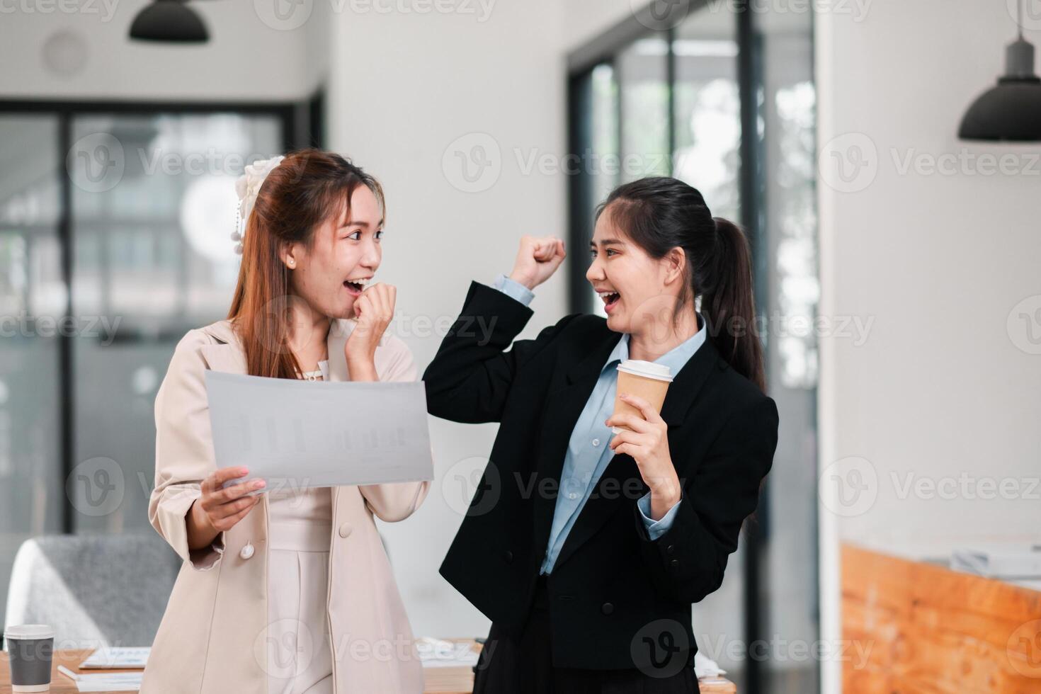 Two businesswomen in a modern office celebrate success with a document and coffee, expressing excitement and teamwork. photo