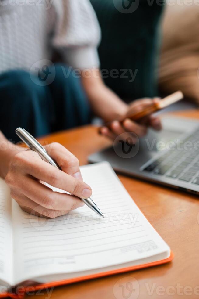 Close-up of a person taking notes in a notebook with a pen, holding a smartphone, and using a laptop on a wooden table. photo