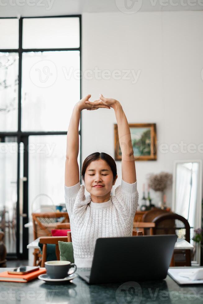Woman enjoying a stretch at her home office desk, surrounded by a cozy and modern interior with a laptop and coffee. photo