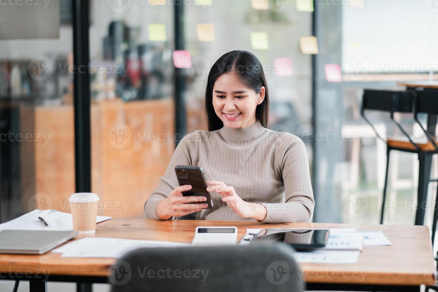 Smiling young woman using smartphone at a desk in a modern office, surrounded by documents and coffee. photo