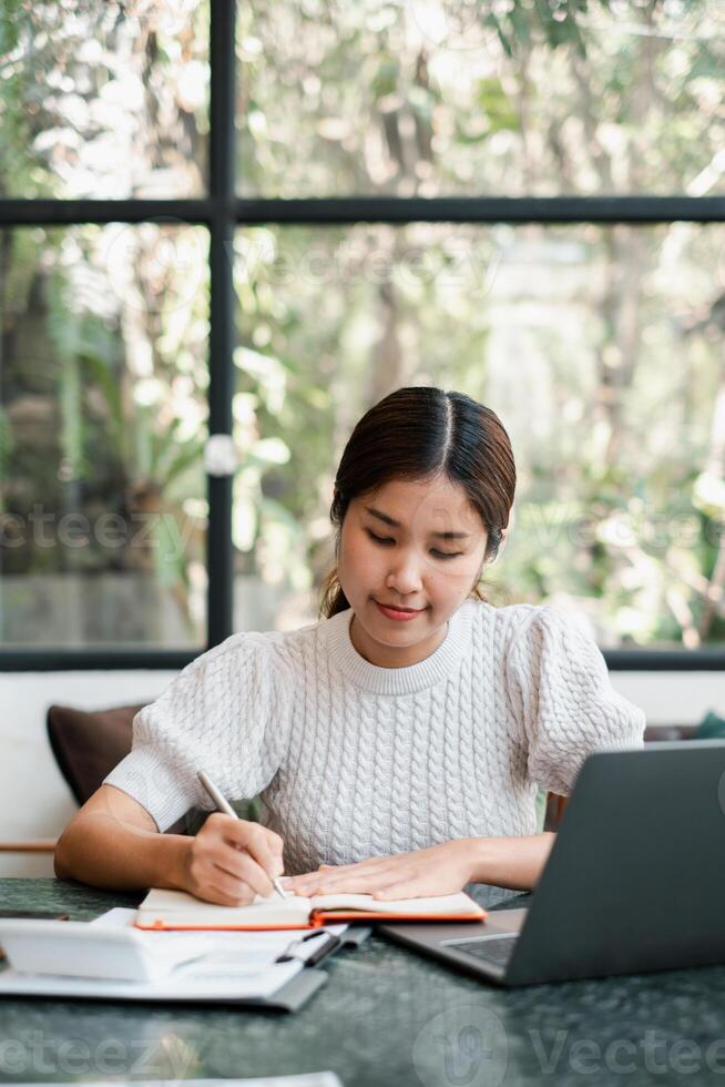 A focused young woman writes in a notebook at a home office desk, with a laptop and natural light streaming through a large window. photo