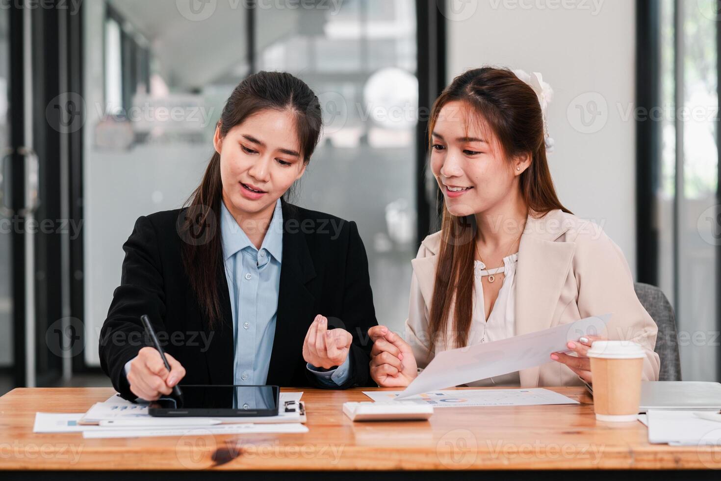 Two businesswomen discussing work at a desk with a tablet and documents in a contemporary office environment. photo