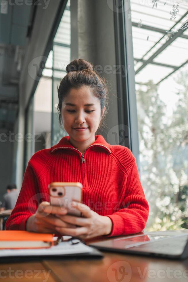 Young woman in a red sweater using her smartphone at a cafe, with natural light from large windows creating a cozy atmosphere. photo