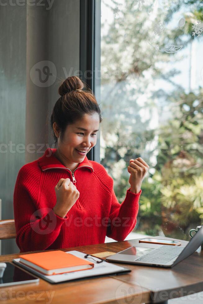A joyful woman in a red sweater celebrates a success while working on her laptop in a bright, modern office space. photo