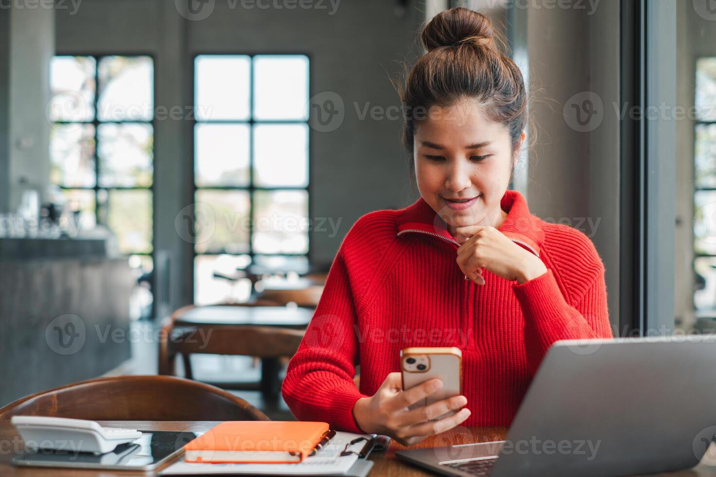 A young woman in a red sweater uses her smartphone and laptop at a cafe table, surrounded by notebooks and a bright, airy atmosphere. photo