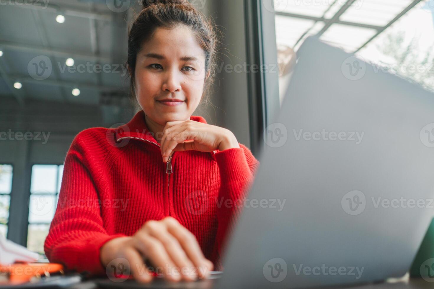 Focused woman in a red sweater working on a laptop in a modern office with natural light. photo