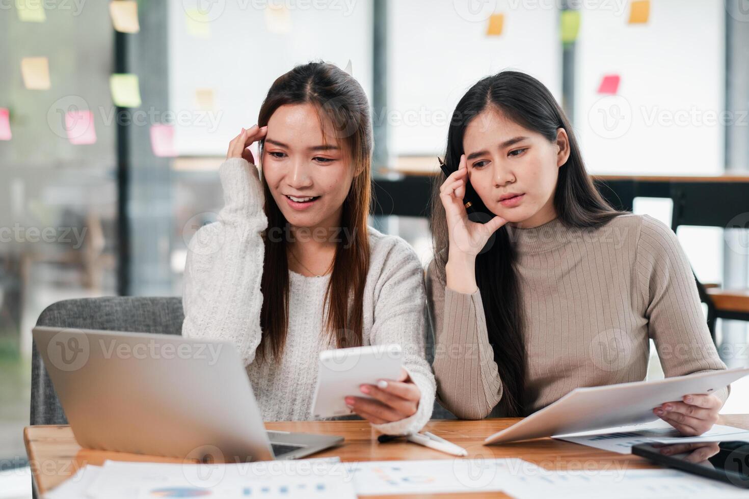 Two young women working together in a modern office, using laptops and documents for a project. photo