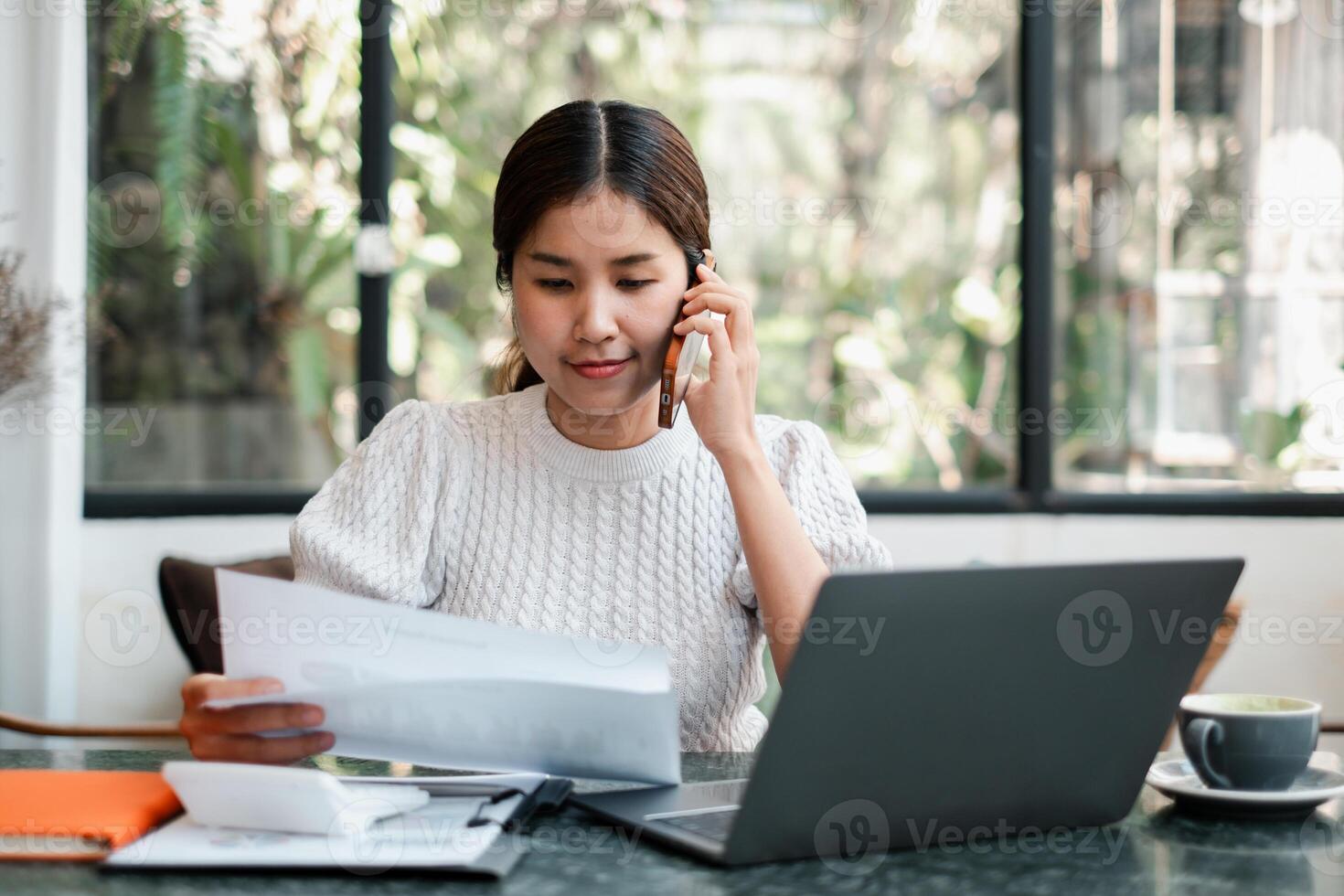 Focused woman in a modern office setting, working on a laptop and talking on the phone, surrounded by documents. photo