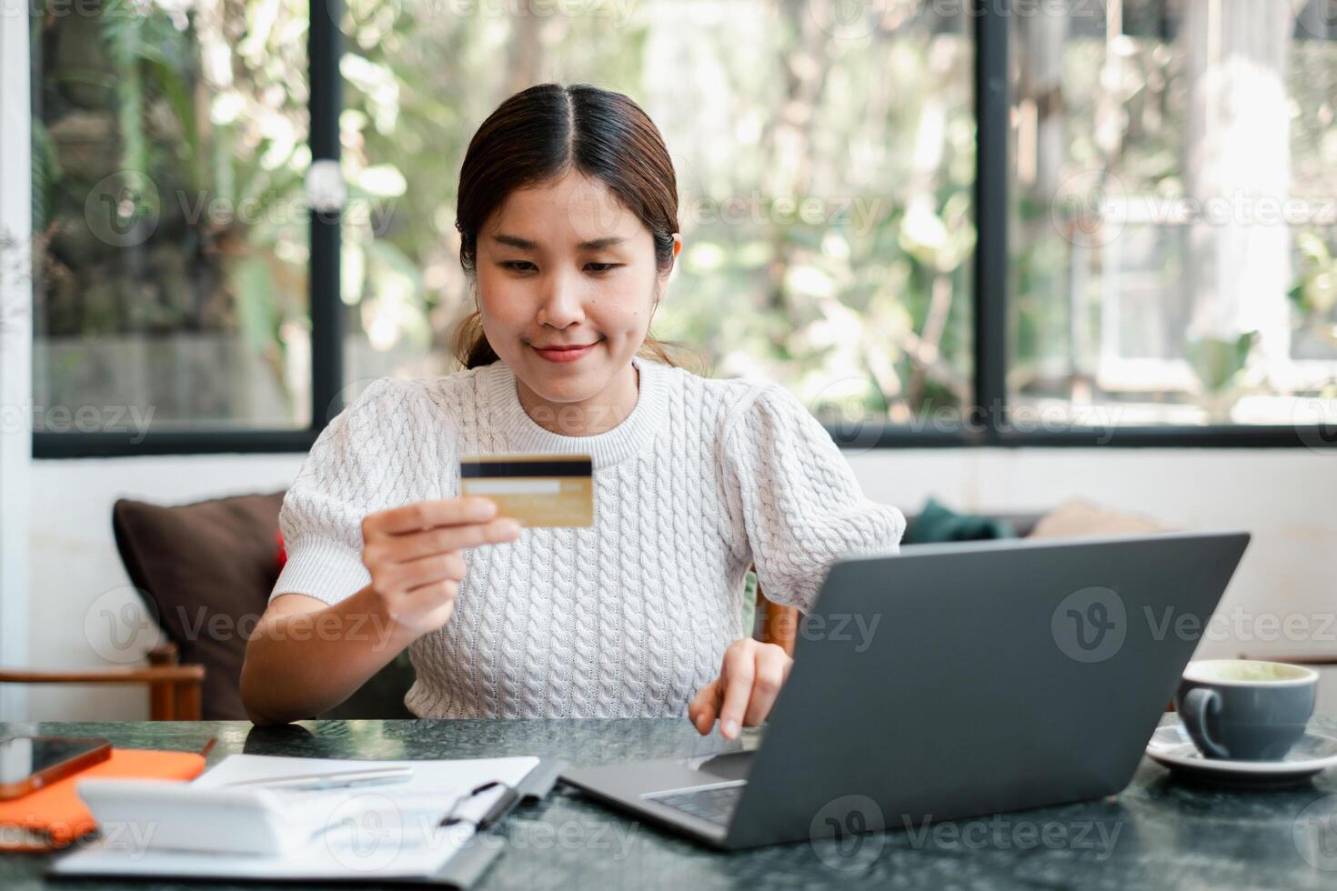 A young woman uses a laptop and credit card for online shopping in a cozy cafe with natural light and greenery. photo