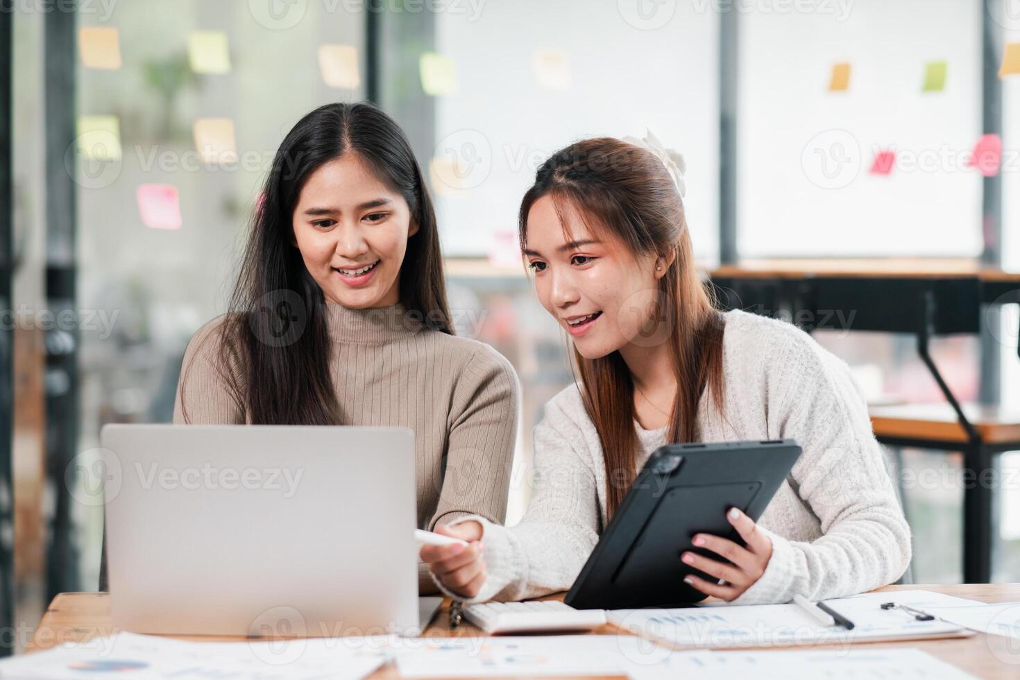 Two young women working together in a modern office, using a laptop and tablet for collaboration and brainstorming. photo