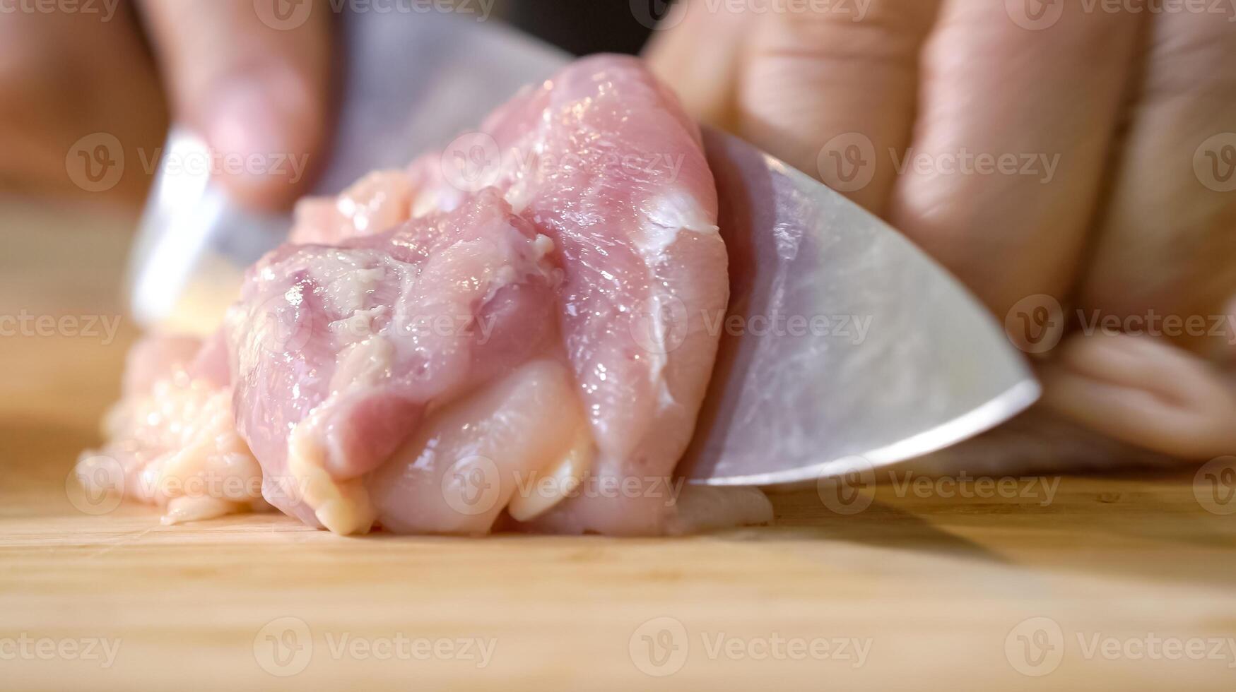 A chef using a kitchen knife slices uncooked chicken meat on a chopping board, the close up view of preparing food ingredients photo
