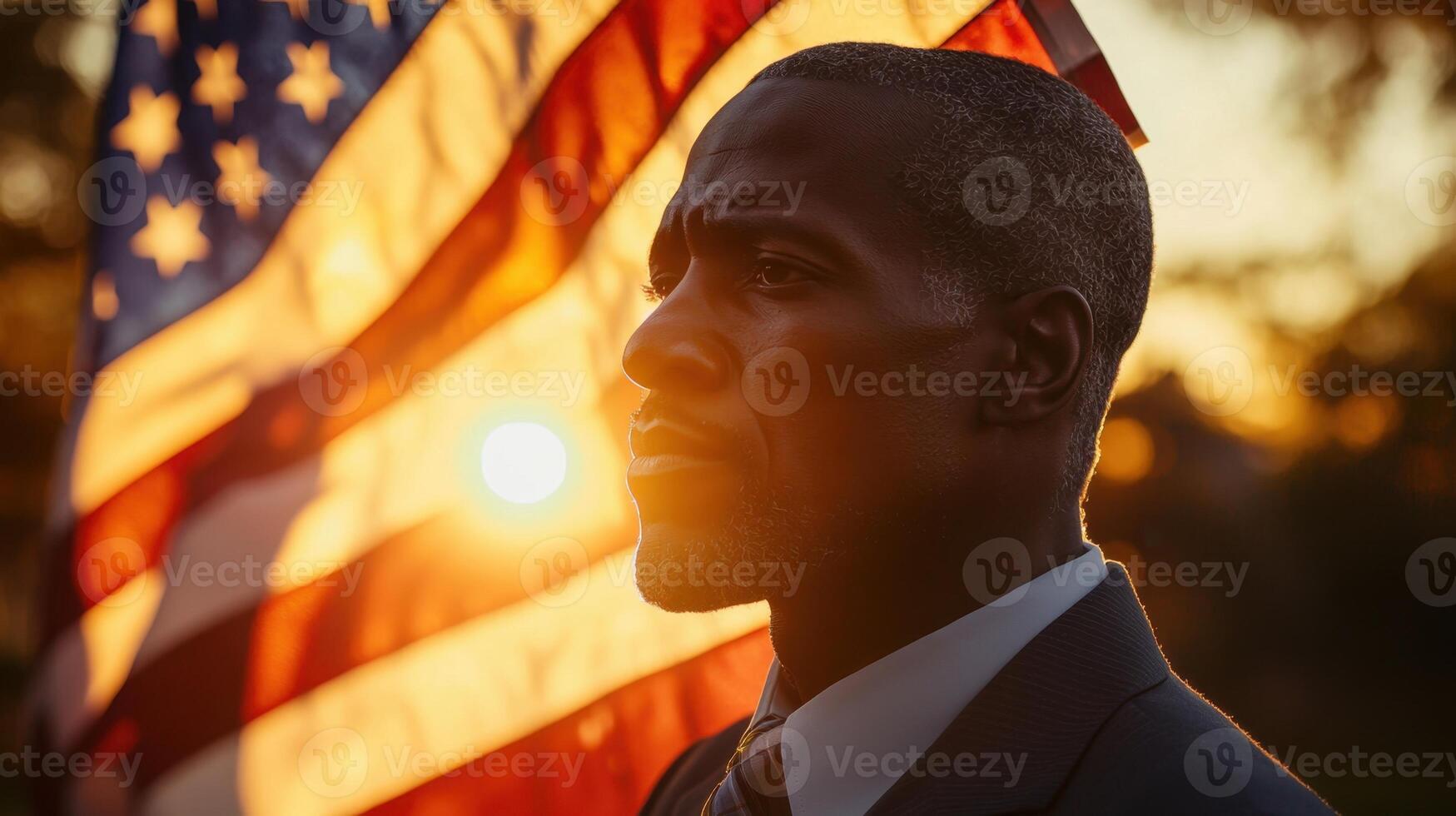 A man stands proudly with a serious expression, silhouetted against a large American flag during a vibrant sunset. The warm glow enhances the patriotic atmosphere photo