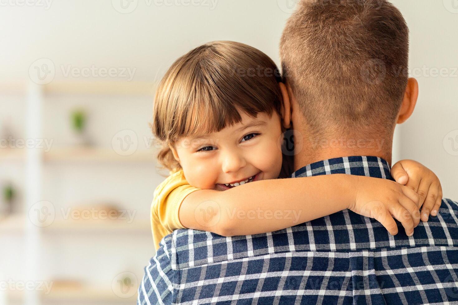 I love my daddy. Close up portrait of happy cute little boy embracing his father and smiling to camera, back view of unrecognizable man photo