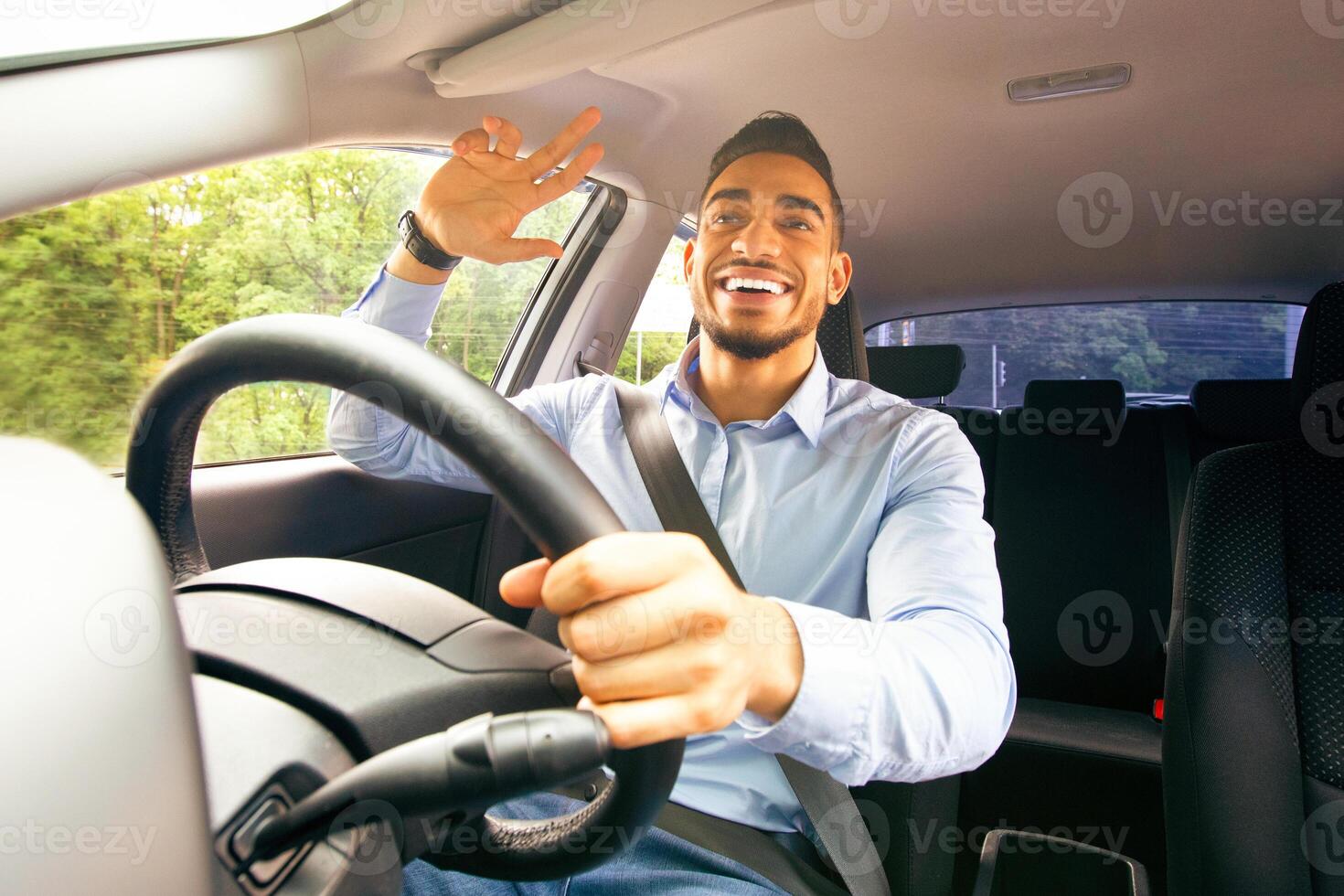 Cheerful middle-eastern guy in formal outfit driving comfortable auto, greeting someone passing by, shot from car dashboard. Handsome businessman going to office by automobile, copy space photo