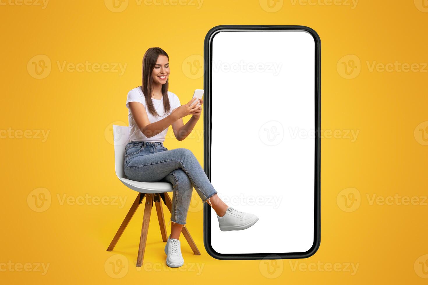 Cheerful smiling female sitting on chair near big cell phone with empty white screen, using mobile device, checking new cool app on orange studio background, mock up for website or application design photo