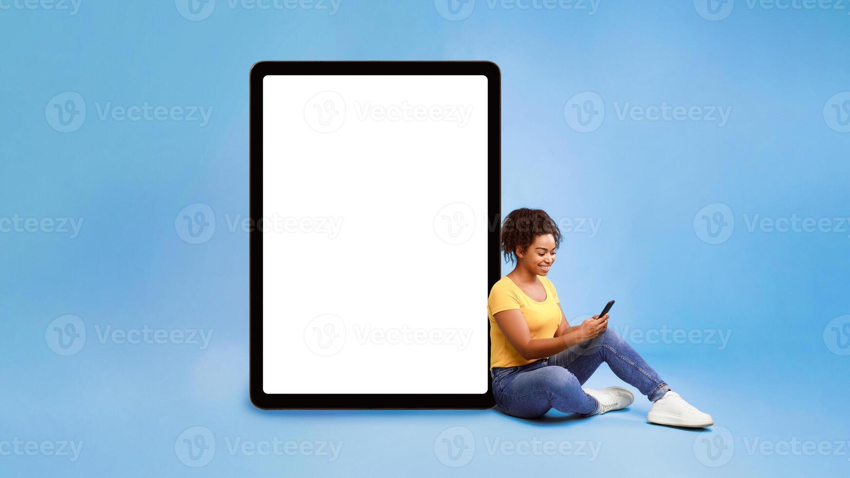 Full length of young African American lady using cellphone, sitting next to giant tablet pc with empty white screen, blue background. Mockup for app or website, space for advertisement design photo