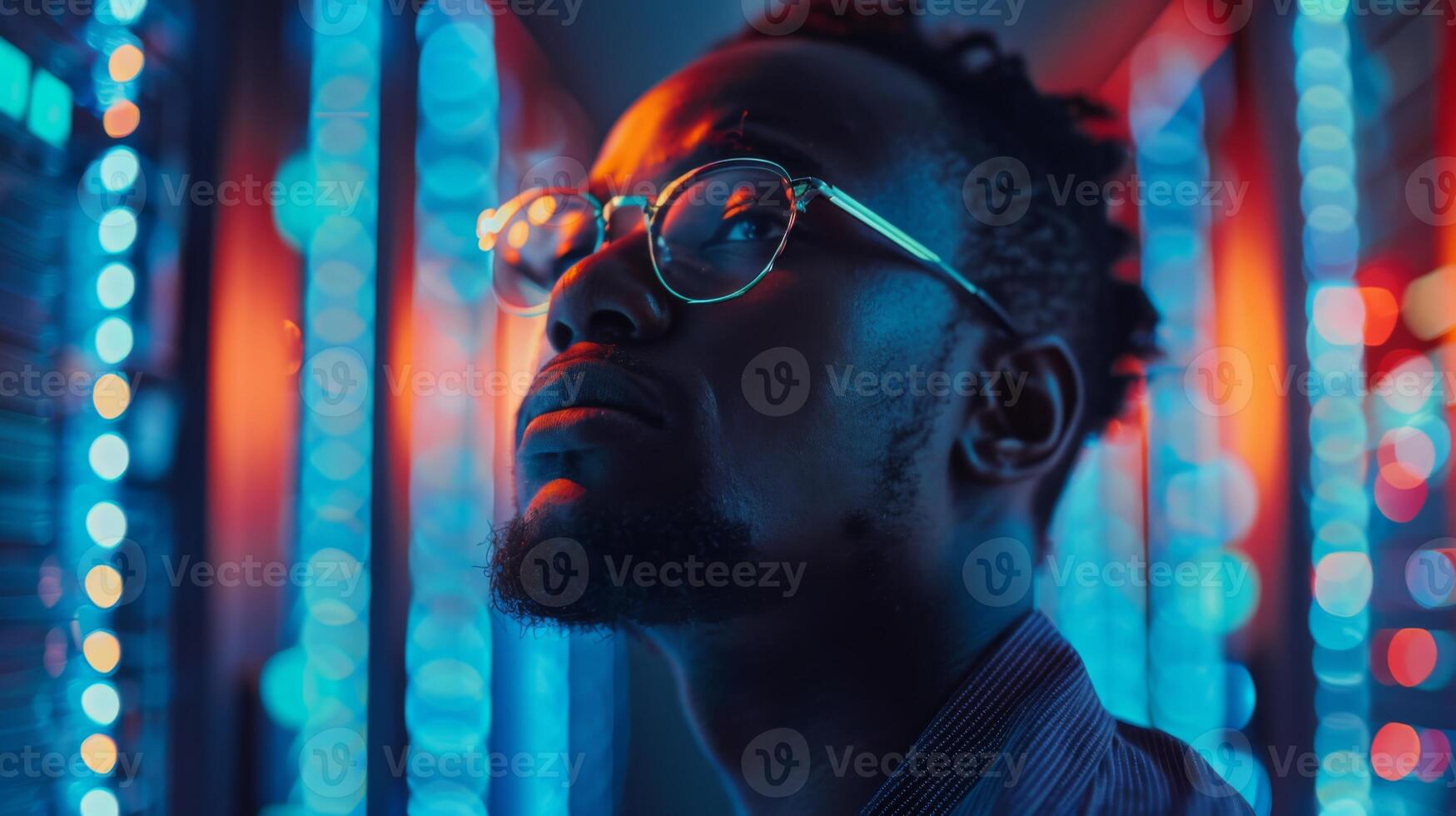 A young man with glasses stands proudly, gazing upward with a serious expression, surrounded by vibrant blue and red lights typical of a modern tech atmosphere during an evening event. photo