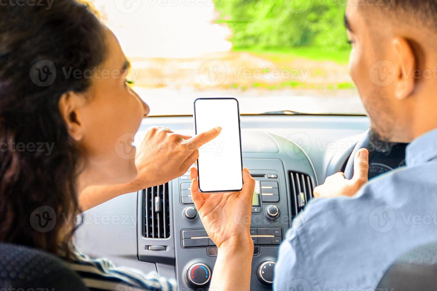 Young mixed race couple having car trip together, using mobile application on smartphone, shot from back seat, closeup. Brunette lady pointing at cellphone with empty screen, mockup photo