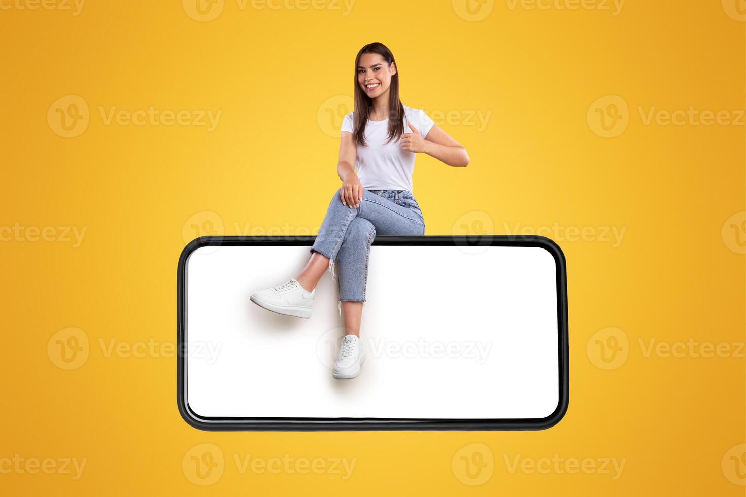 Positive casual millennial lady sitting on big horizontal smartphone with blank screen and showing thumbs up ok gesture, recommending newest mobile application, isolated on orange studio background photo