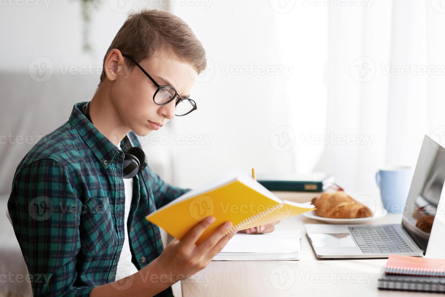 Side view of smart schooler reading book and using laptop, working on project at home, free space photo
