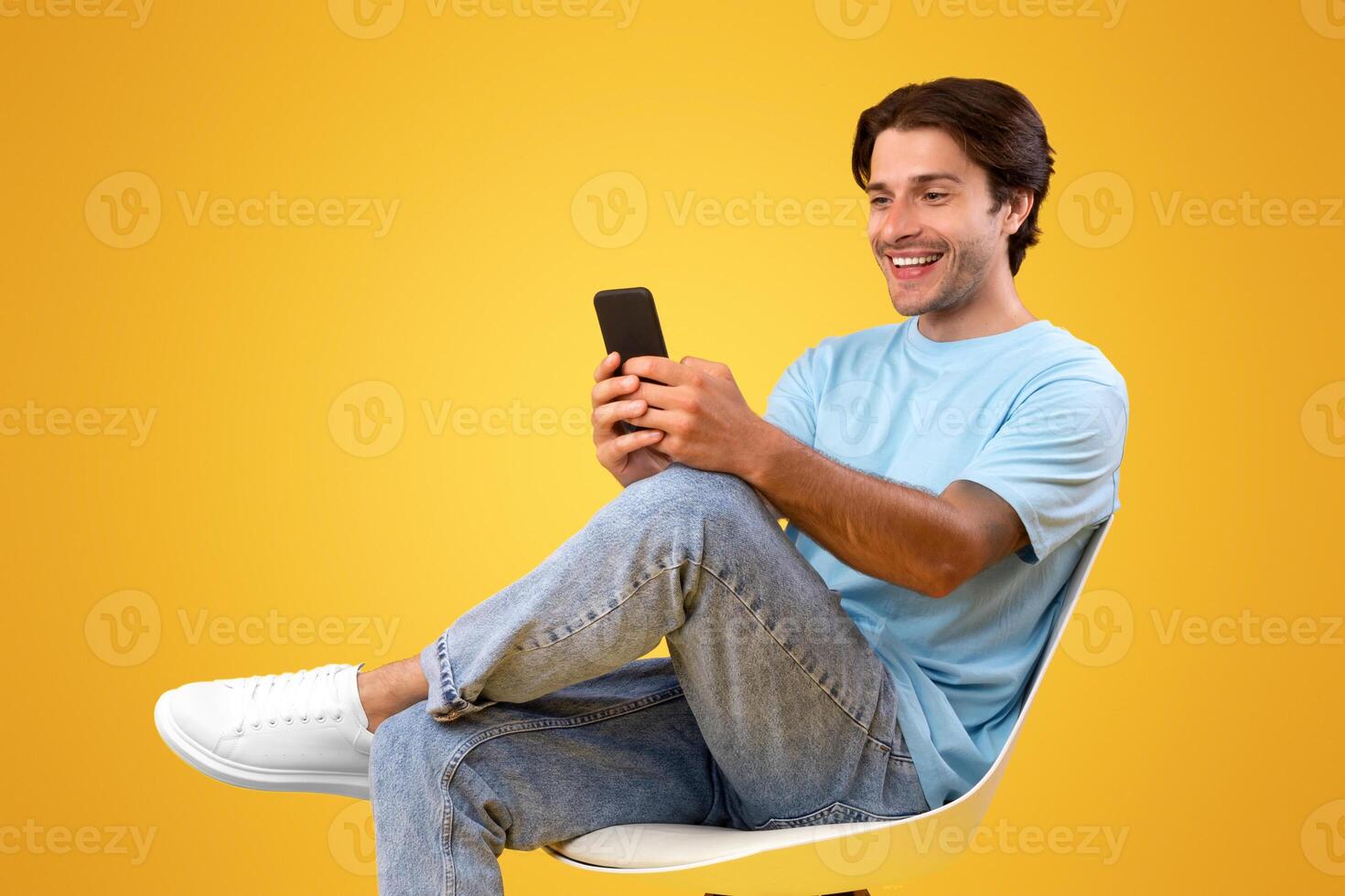 People And Technology Concept. Portrait of smiling young man using smartphone sitting on chair isolated on orange studio background. Excited casual guy chatting online, browsing social media photo