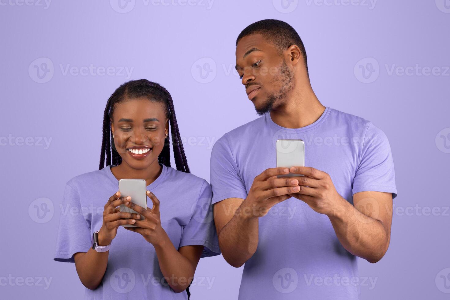 Portrait of curious black guy trying to look at his girlfriend's smartphone, smiling lady holding using her gadget and chatting, couple standing isolated on violet purple background. Distrust Concept photo