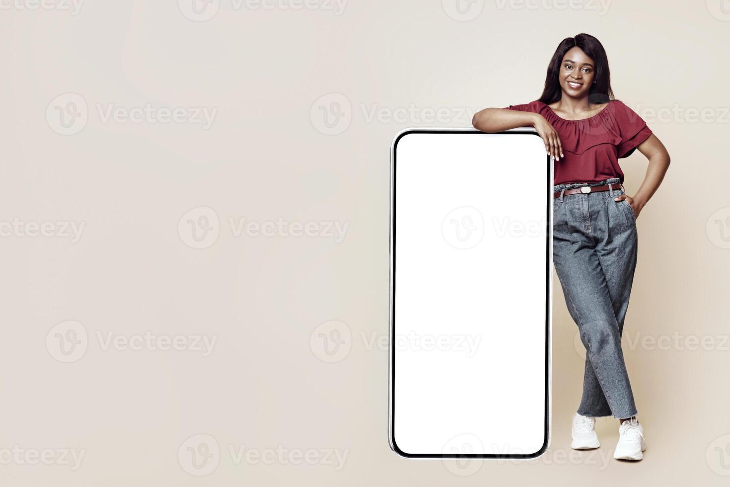 Pretty young black woman in casual outfit standing by huge modern smartphone with white empty screen and smiling at camera, mockup, copy space, beige studio background. Modern technologies concept photo