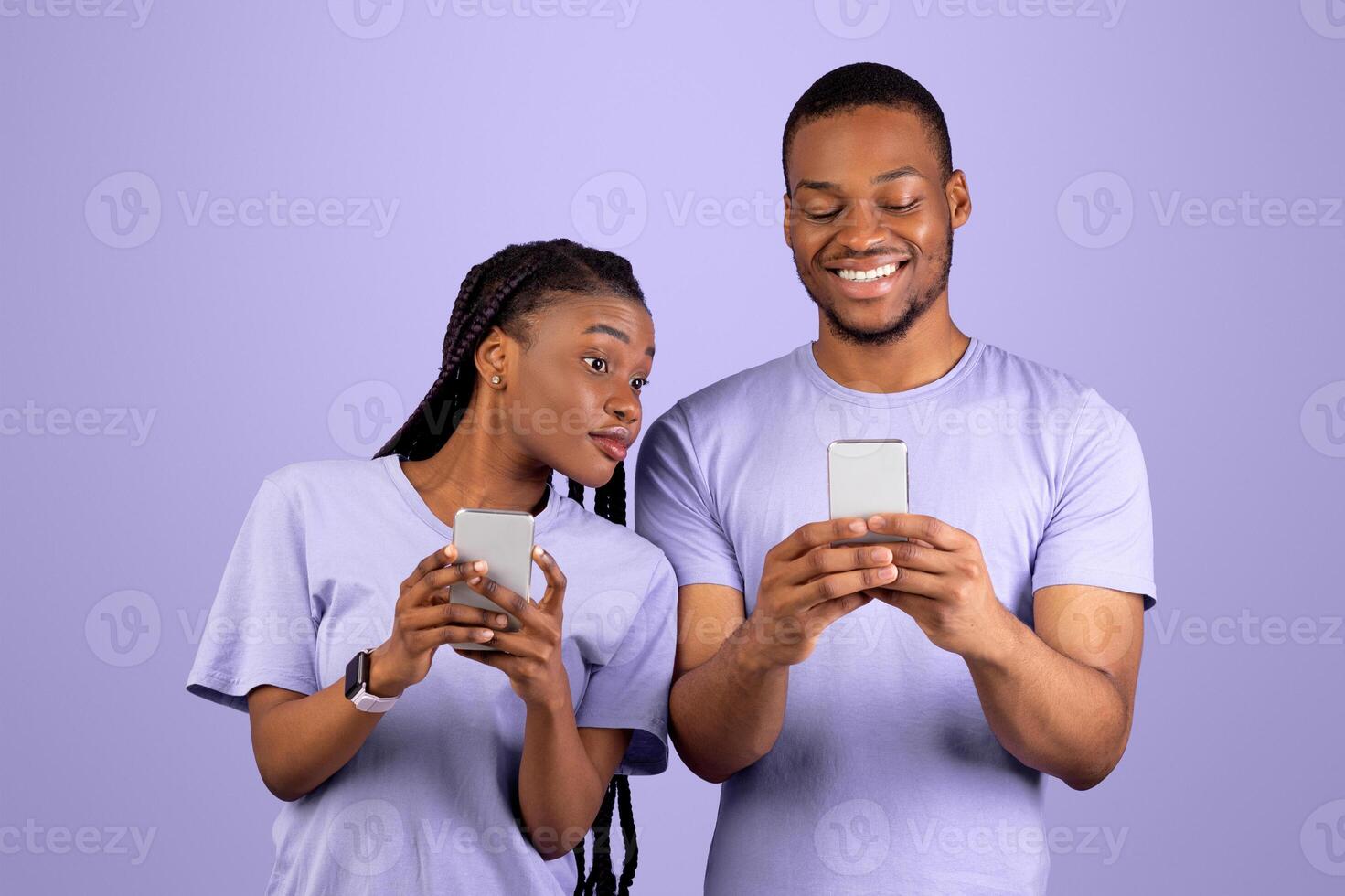 Portrait of curious black woman trying to look at her boyfriend's smartphone, smiling man holding using his gadget and chatting, couple standing isolated on violet studio background. Distrust Concept photo