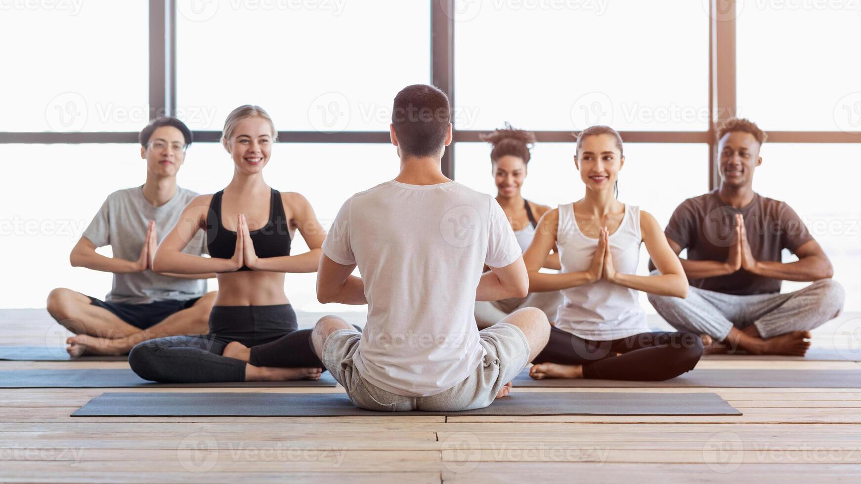 International group of young people attending yoga class in modern studio, meditating with coach in lotus position photo