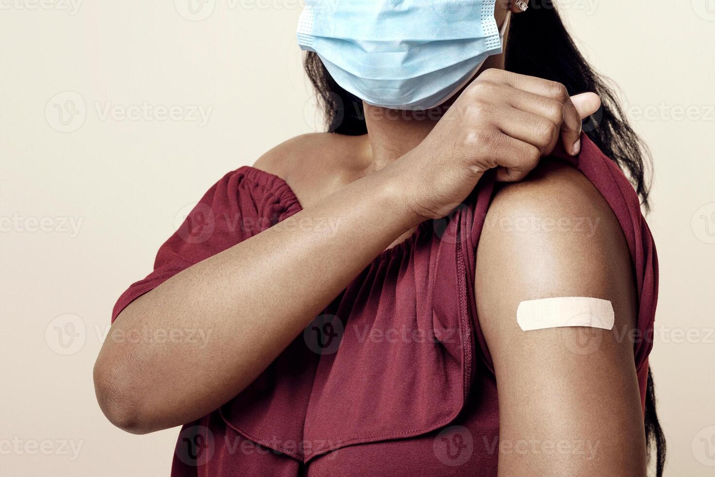 Portrait of millennial black woman with adhesive medical band on her shoulder wearing protective face mask, young people got vaccinated while COVID-19 pandemic, studio background, isolated photo