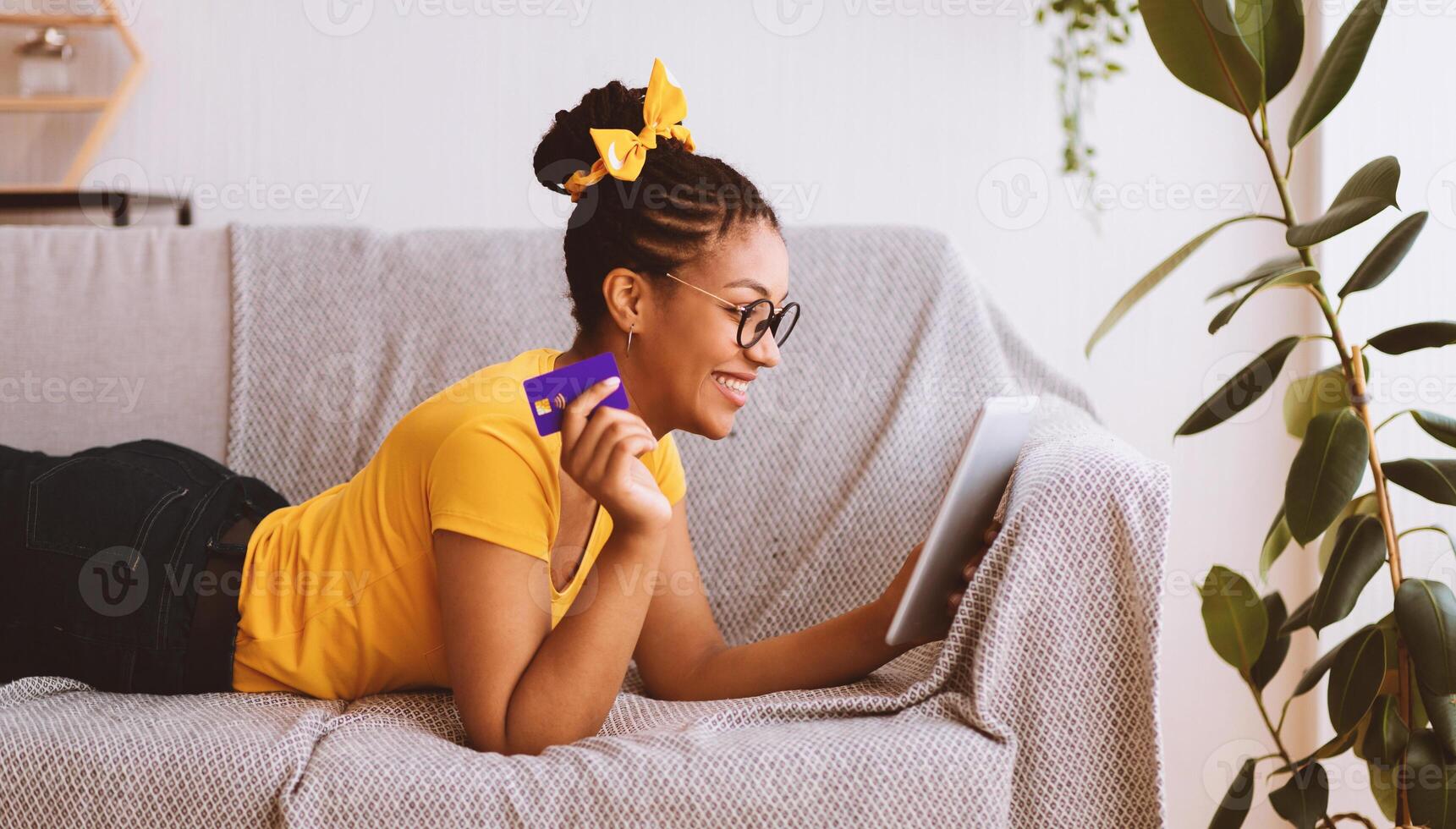 Online Shopping Concept. Happy Black Woman In Glasses Holding And Using Digital Tablet And Credit Card, Buying And Ordering Things In Internet Shop Lying On The Couch. Sale, Retail And Purchase photo