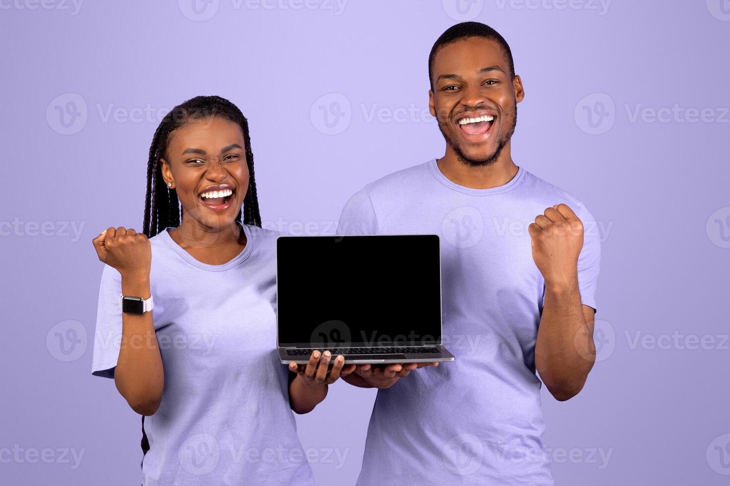 Yes. Portrait of excited happy black couple holding pc laptop with blank screen shaking clenched fists making winner gesture, showing device with empty free space for mock up, violet studio background photo