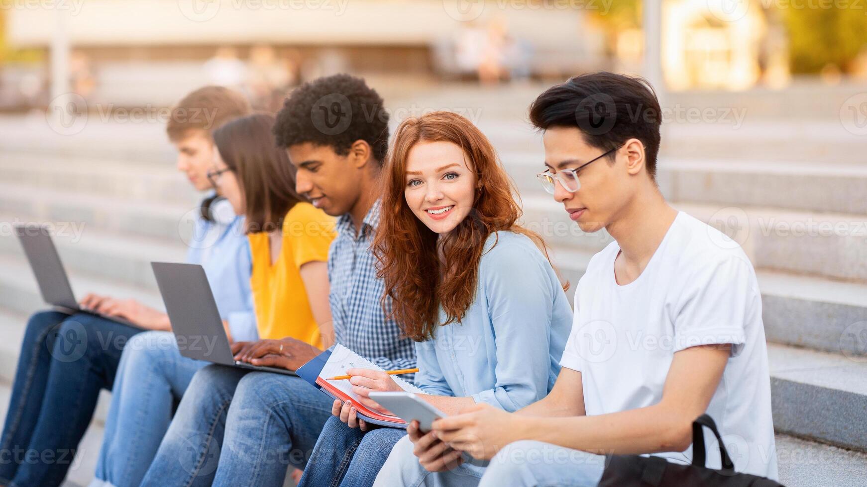Group Of Students Learning Using Gadgets Sitting On College Steps Outdoor Preparing For End-Of-Semester Exams Together. photo