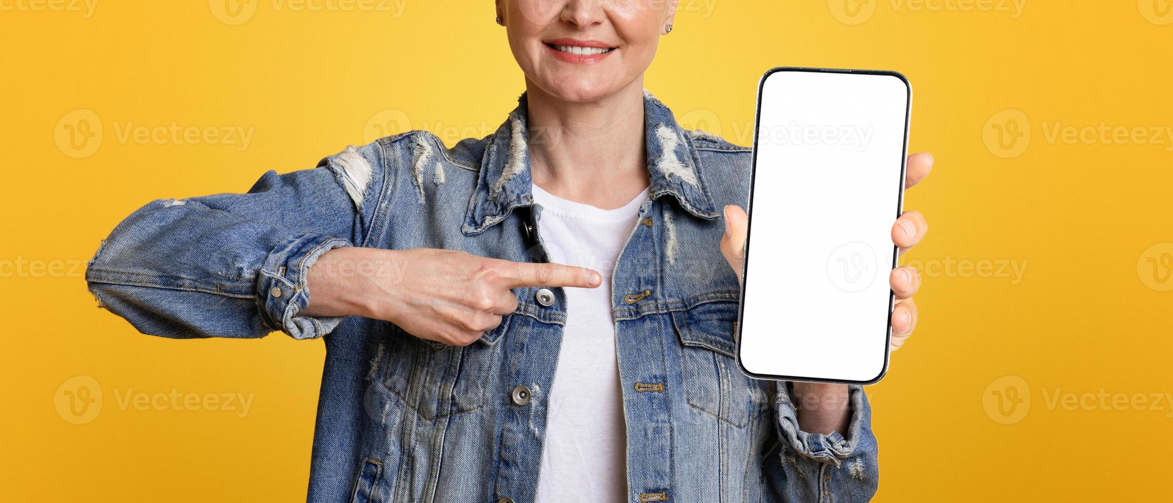 Smartphone Mockup. Smiling Middle Aged Woman Pointing At Black Blank Mobile Screen, Standing Over Yellow Background, Cropped Image, Panorama photo