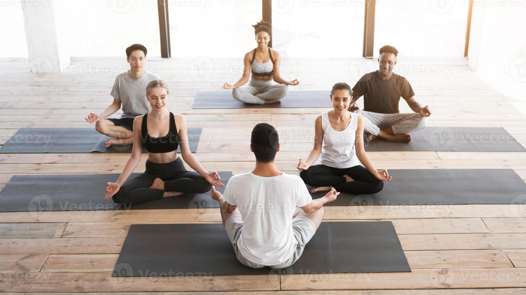 Group of young people having yoga class with coach in studio, sitting in lotus position and smiling, copy space photo