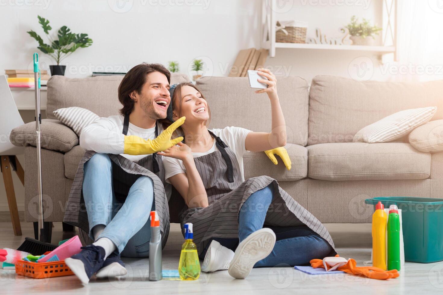 Quarantine Pass Time. Smiling young couple taking selfie on smartphone after spring-cleaning apartment, sitting on floor, free space photo