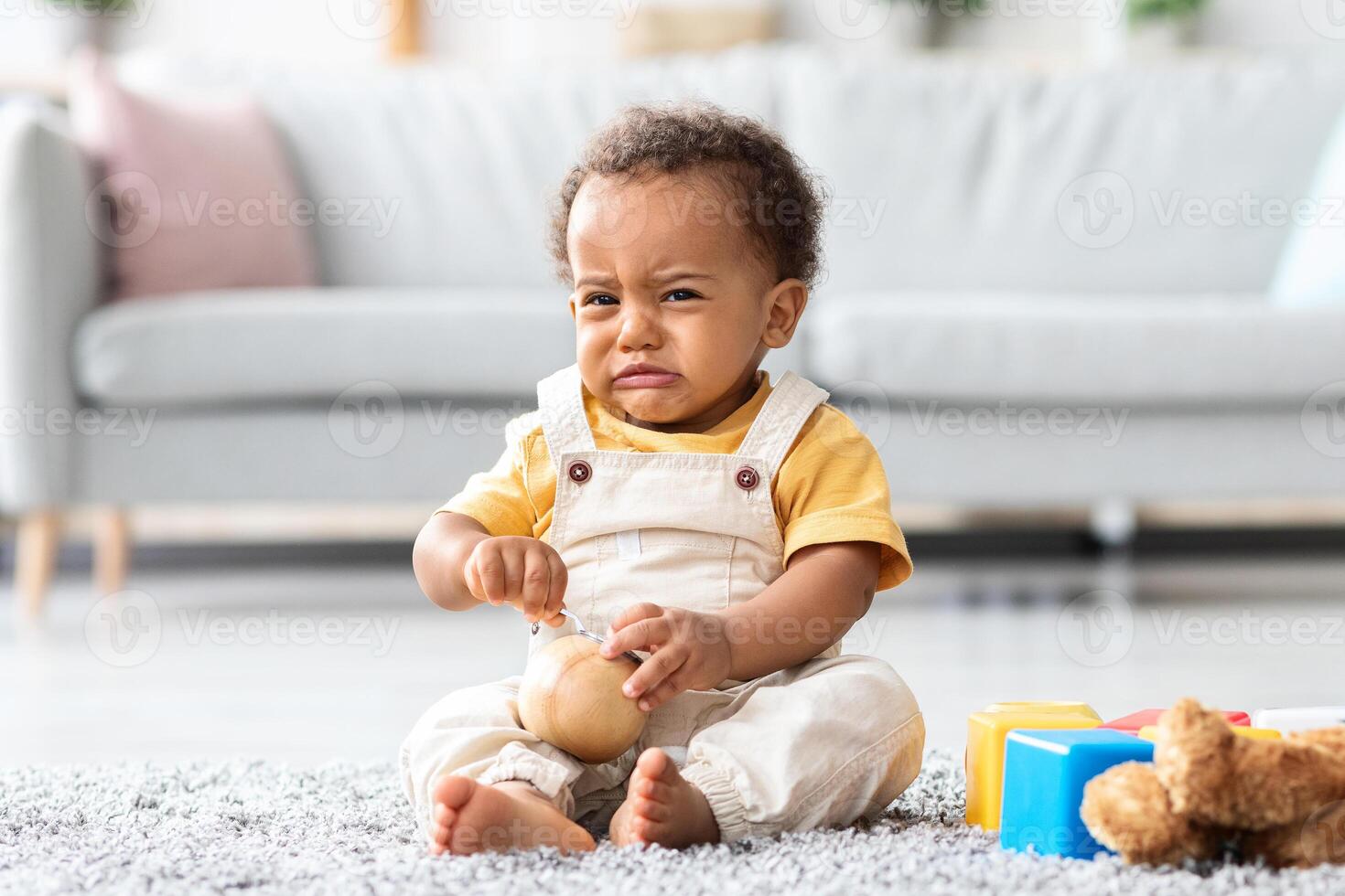 Portrait Of Crying Cute African American Infant Boy Sitting On Floor In Living Room, Upset Toddler Child Wearing Jumpsuit Whining And Sobbing At Home, Feeling Sad And Lonely, Copy Space photo