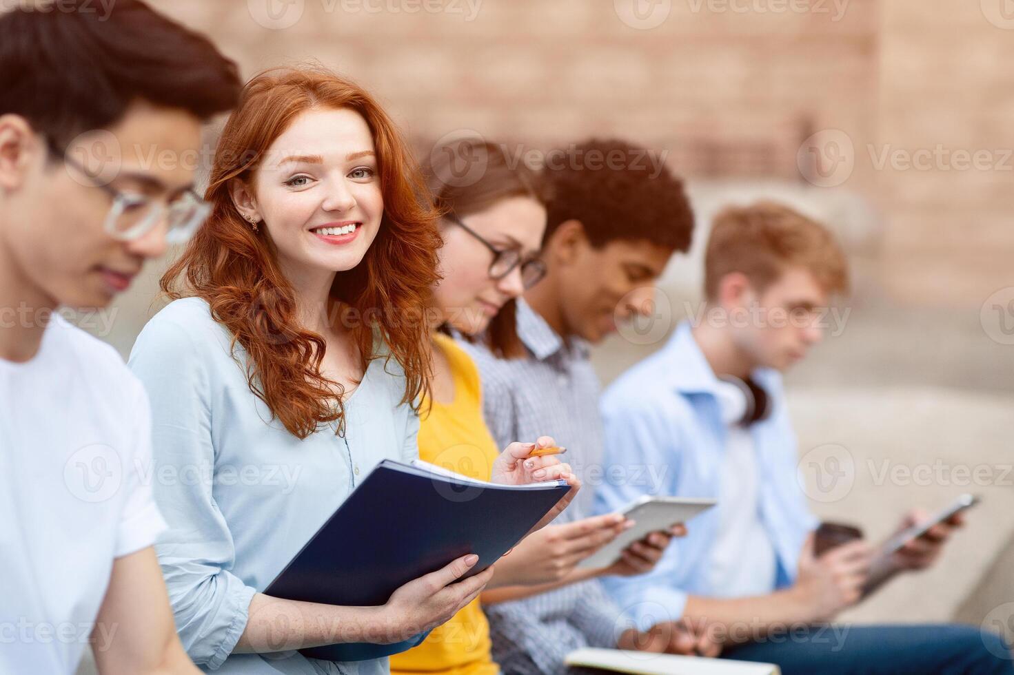 Learning Concept. Portrait of cute red-haired girl studying outdoors with her friends and looking at camera photo