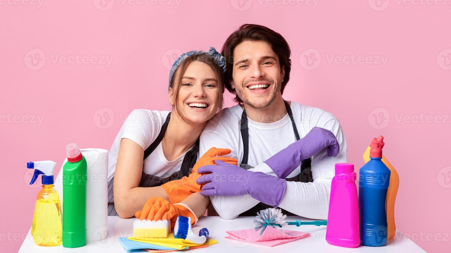Cheerful Couple Sitting At Table With Lots Of Detergents And Cleaning Supplies, Hugging And Smiling At Camera, Ready To Spring-Clean, Pink Background photo
