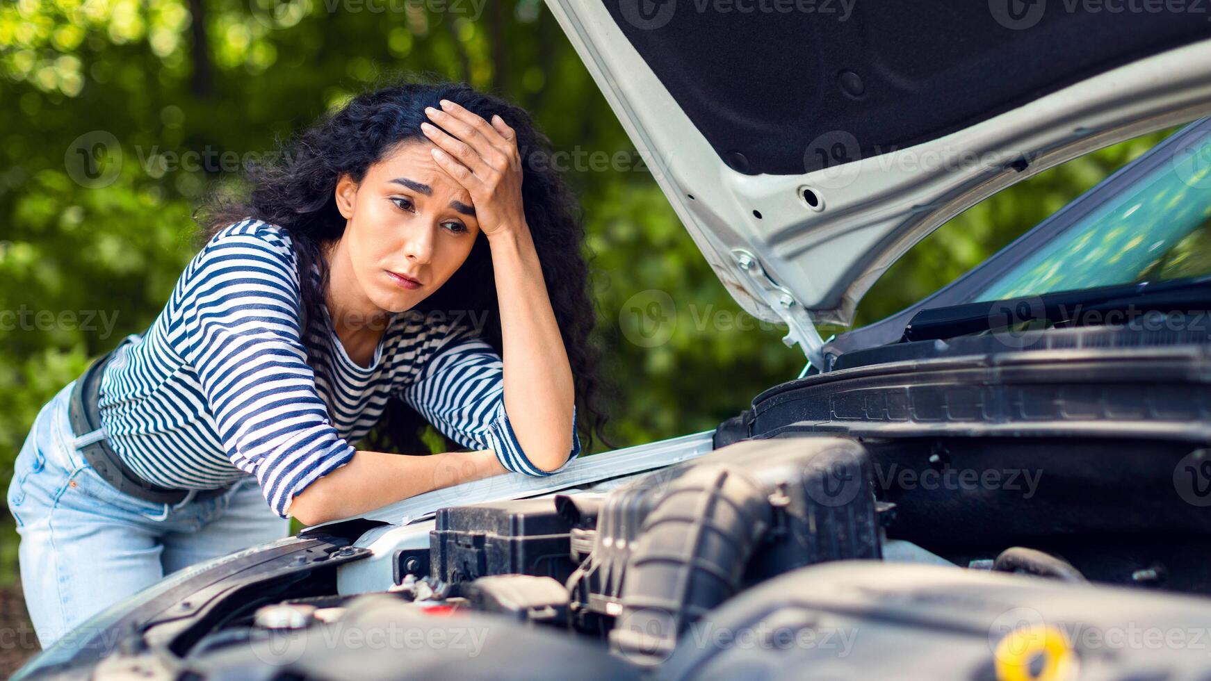 Sad brunette woman leaning on car hood, experiencing broken engine while car trip, waiting for auto service, closeup. Depressed young lady having troubles with her car, waiting for help photo
