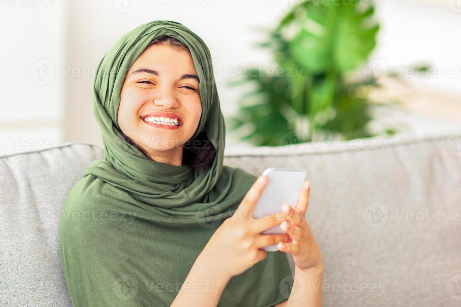 Smiling Muslim adolescent girl in hijab chatting online, sitting on couch with smartphone at home. Indian teenager using mobile device, communicating on web, browsing social media photo