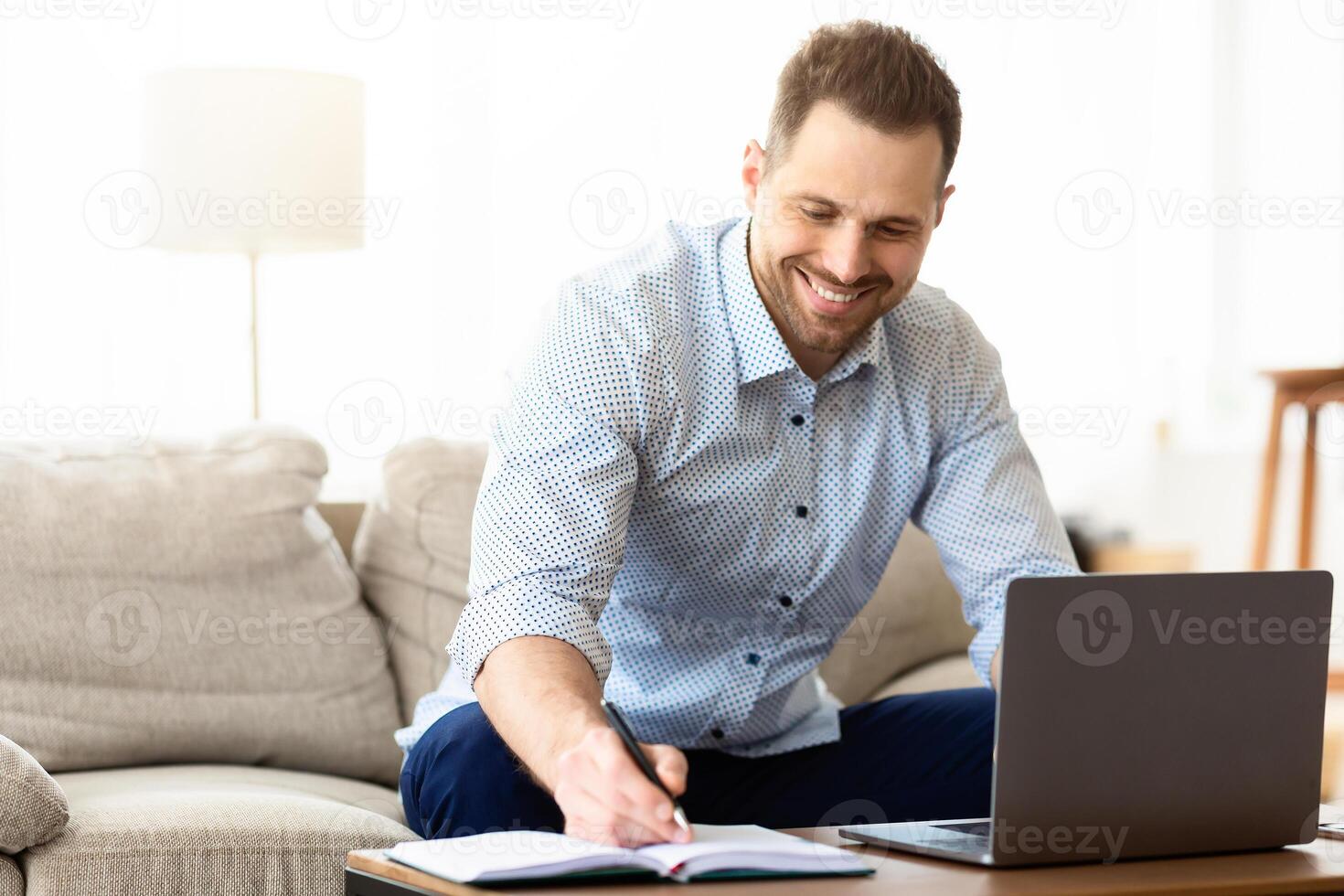 Note Taking Concept. Smiling handsome man using laptop, writing in notebook, sitting on couch, copy space photo
