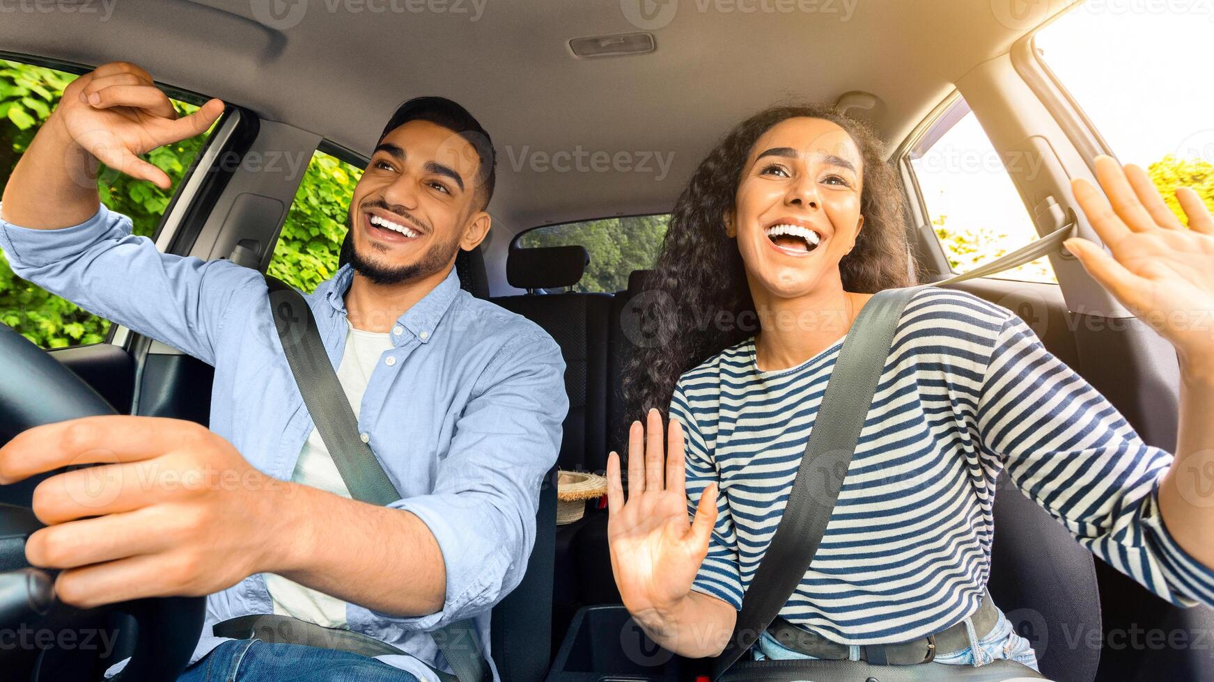 Emotional millennial mixed race couple handsome arab guy and pretty brunette lady having fun while car trip, listening to music and cherfully singing, shot from inside the auto, sun flare photo
