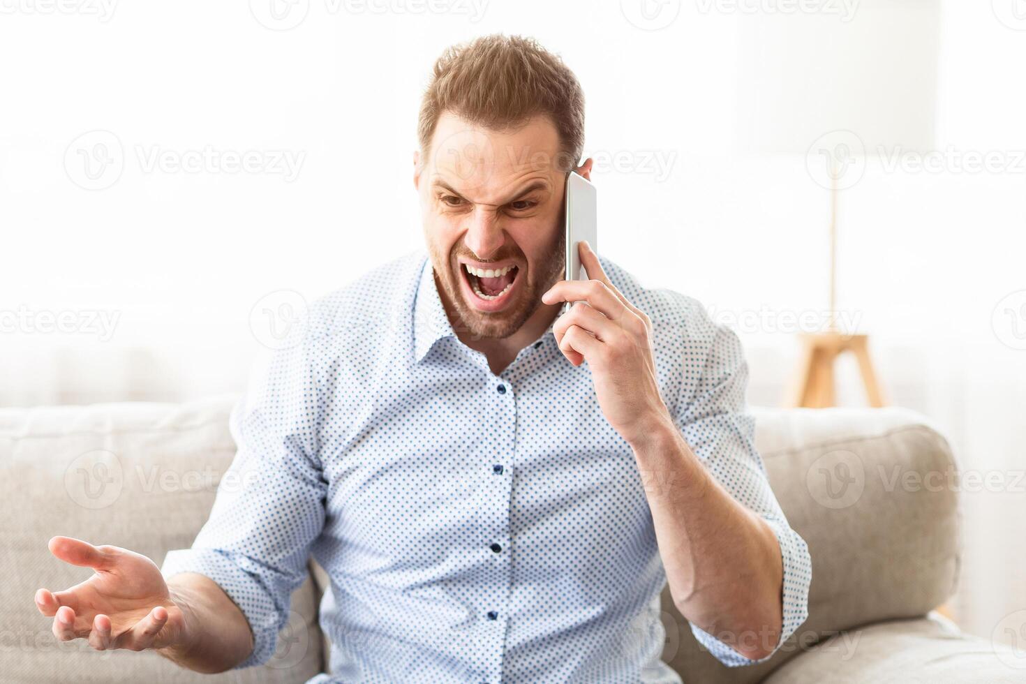 Angry irritated man talking on mobile phone and shouting, sitting on sofa, copyspace photo