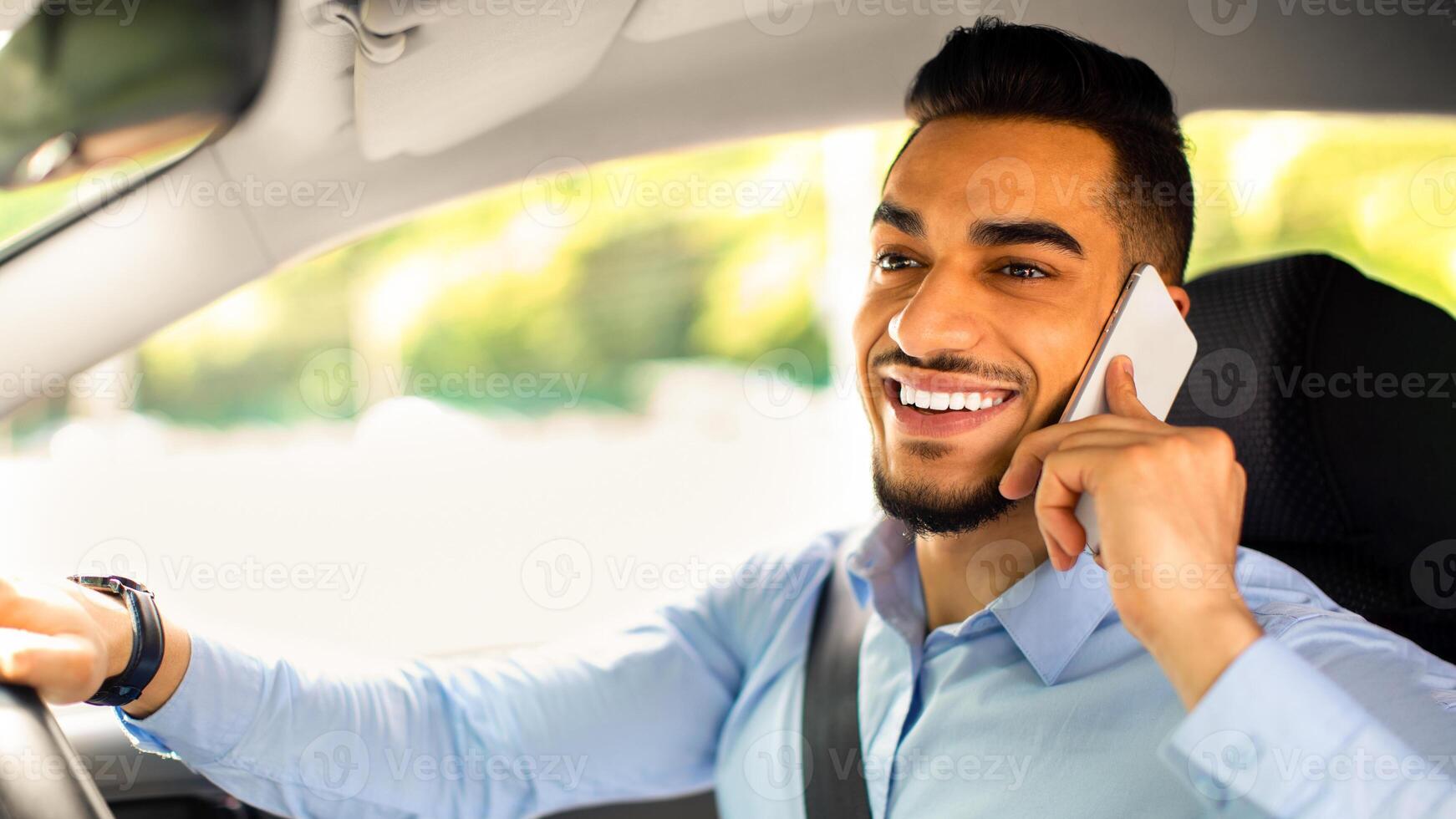 Bad driving habits, unsafe driving, driving and gadgets concept. Cheerful arab man entrepreneur going to office by auto, talking on phone while driving car, side view, copy space, closeup photo