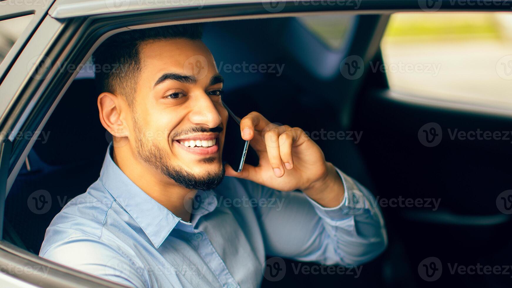 Cheerful middle-eastern young man manager talking to business partner on cellphone while going to office by taxi, sitting at car back seat, closeup portrait, copy space. Businessman lifestyle concept photo