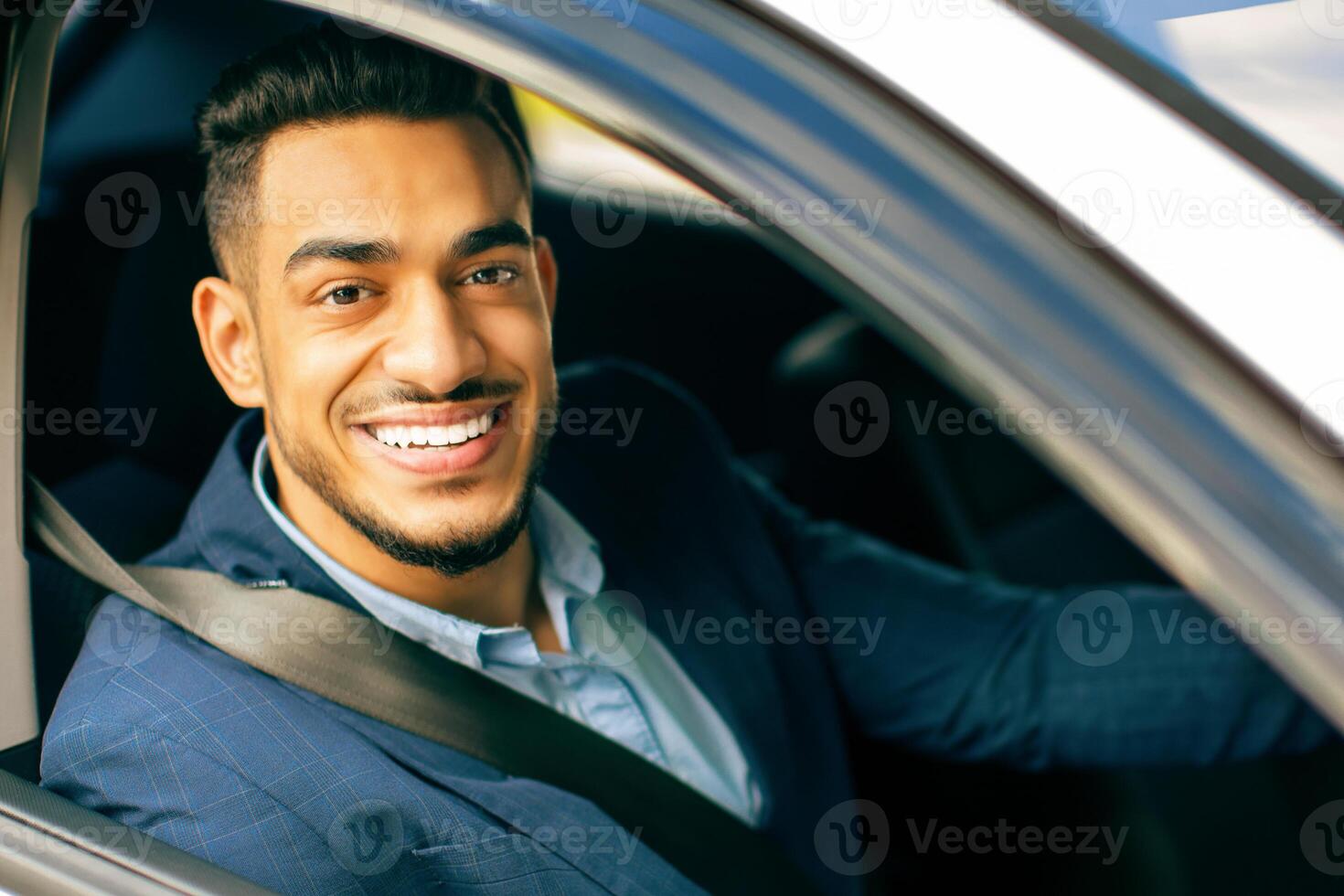 Portrait of smiling middle-eastern young businessman driving car, going to office, shot from outside, closeup. Happy successful arab guy in suit manager going home after good day photo